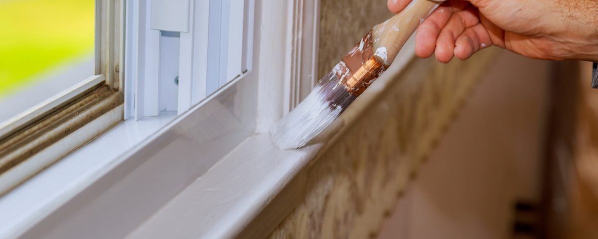 Closeup image of a mans hand with paintbrush while painting window trim