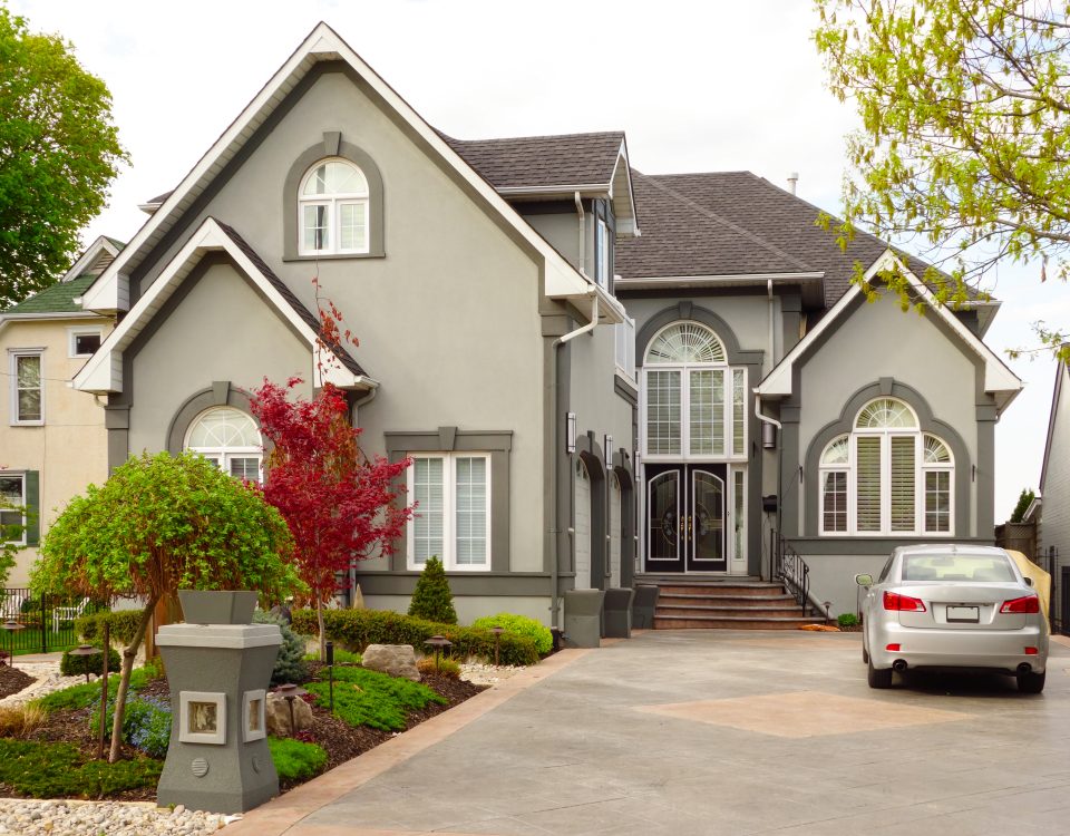 Front view of a large gray house with steep gables, arched windows, and a landscaped yard with a parked car in the driveway.