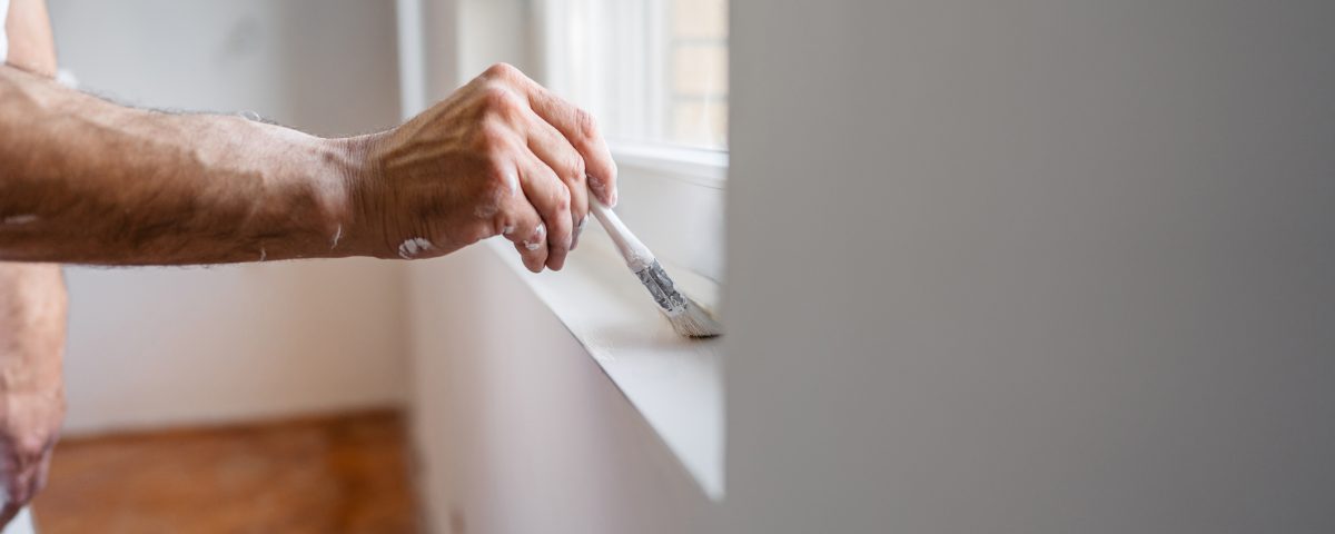 Mature Worker Painting The Window Sill With A Paint Brush In An Empty Apartment Under Renovation - Dow Brothers Painting LTD Close-up of a person painting a windowsill with a brush, with paint on their arm and white shirt sleeve visible.