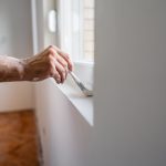 Mature Worker Painting The Window Sill With A Paint Brush In An Empty Apartment Under Renovation - Dow Brothers Painting LTD Close-up of a person painting a windowsill with a brush, with paint on their arm and white shirt sleeve visible.