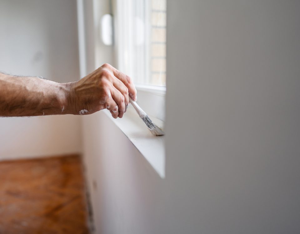 Close-up of a person painting a windowsill with a brush, with paint on their arm and white shirt sleeve visible.